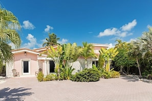 A two-story building with a pink facade, surrounded by tropical vegetation and a paved driveway.