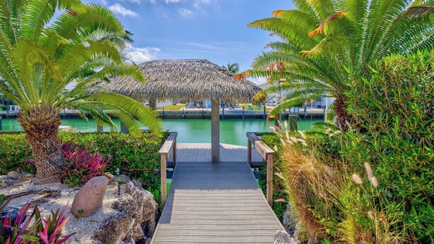 A wooden deck leading to a thatched-roof structure by a body of water, surrounded by tropical plants. - P34 - Beautiful 4 bedroom 2 bath, single family home with private pool and deep water dockage. (Key Colony Beach)