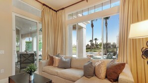 A living room with a beige sectional sofa, a dark wooden coffee table, and a view of a pool and palm trees through large windows. - FALL FLASH SALE -Luxe Pool, Misters Firepit Lounge & Billiards (La Quinta)