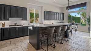 A modern kitchen with dark cabinets, a large island with bar stools, and a view of the outdoors. - FALL FLASH SALE -Luxe Pool, Misters Firepit Lounge & Billiards (La Quinta)