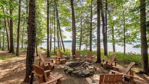 Ein Lagerfeuer, umringt von Holz-Adirondack-Stühlen, in einer bewaldeten Gegend mit Blick auf einen See.
