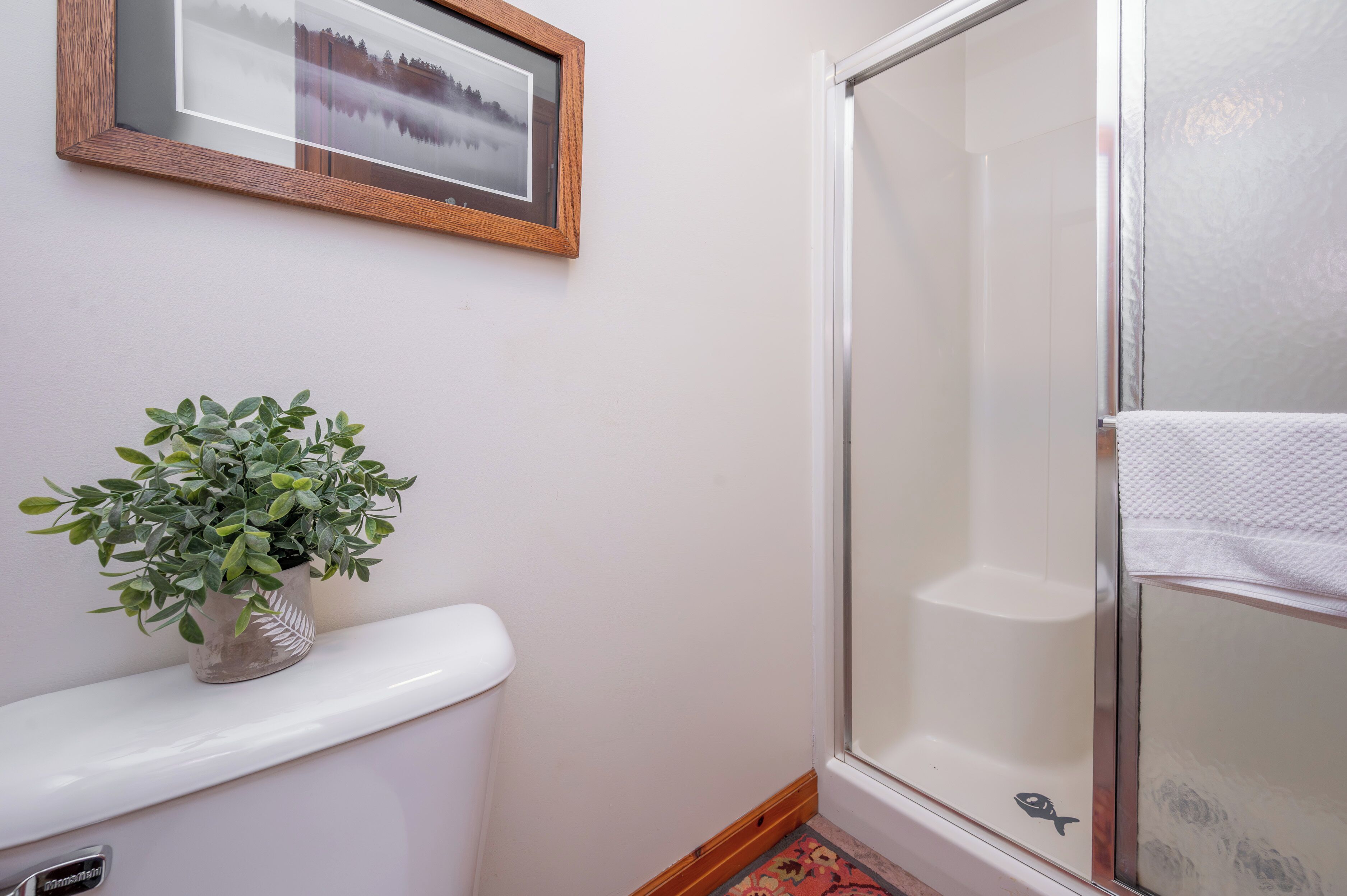 Une salle de bain avec des toilettes blanches, une plante en pot, un tableau encadré au mur et une cabine de douche en verre.