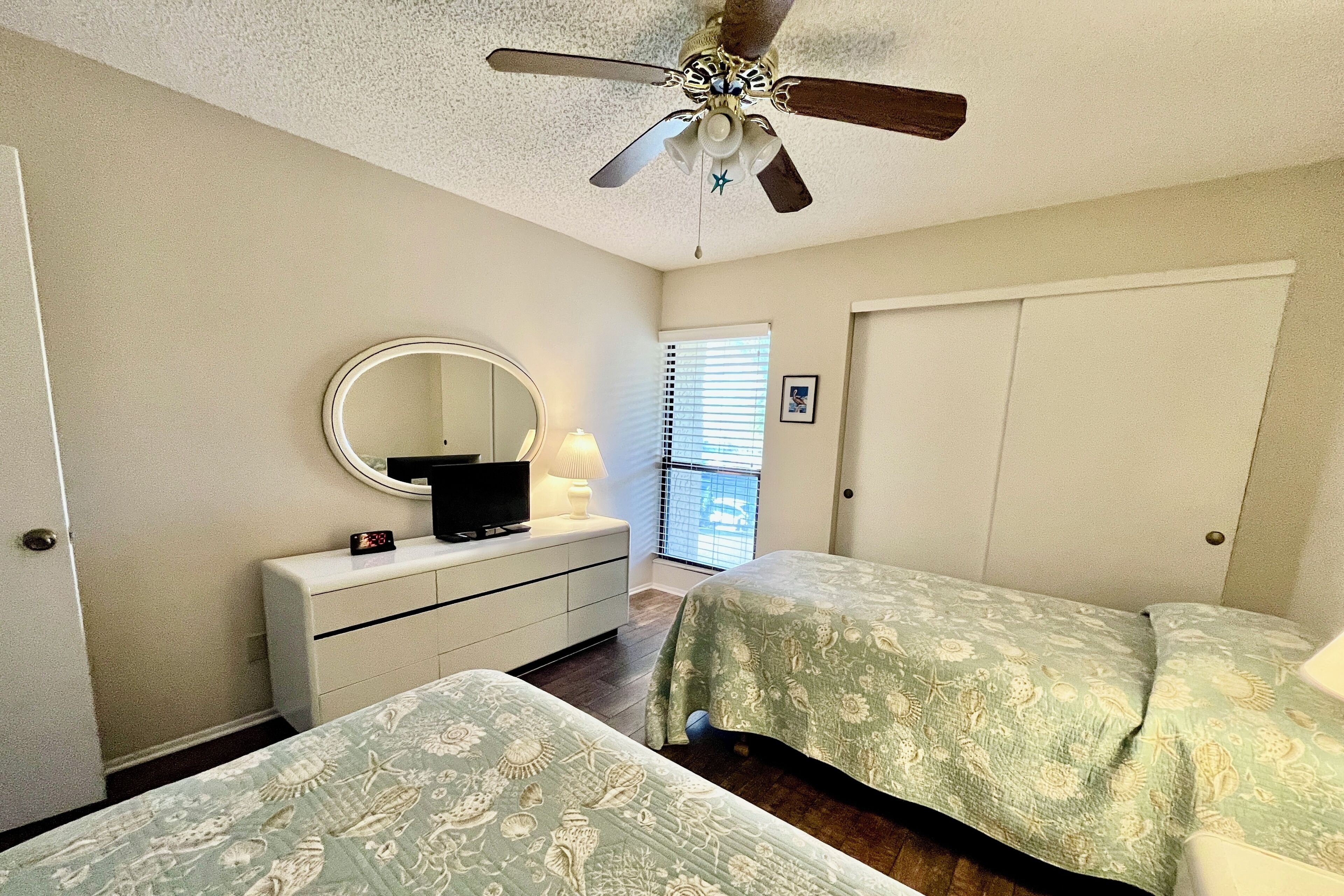 A bedroom with two beds, a ceiling fan, a dresser with a mirror, and a window with blinds.
