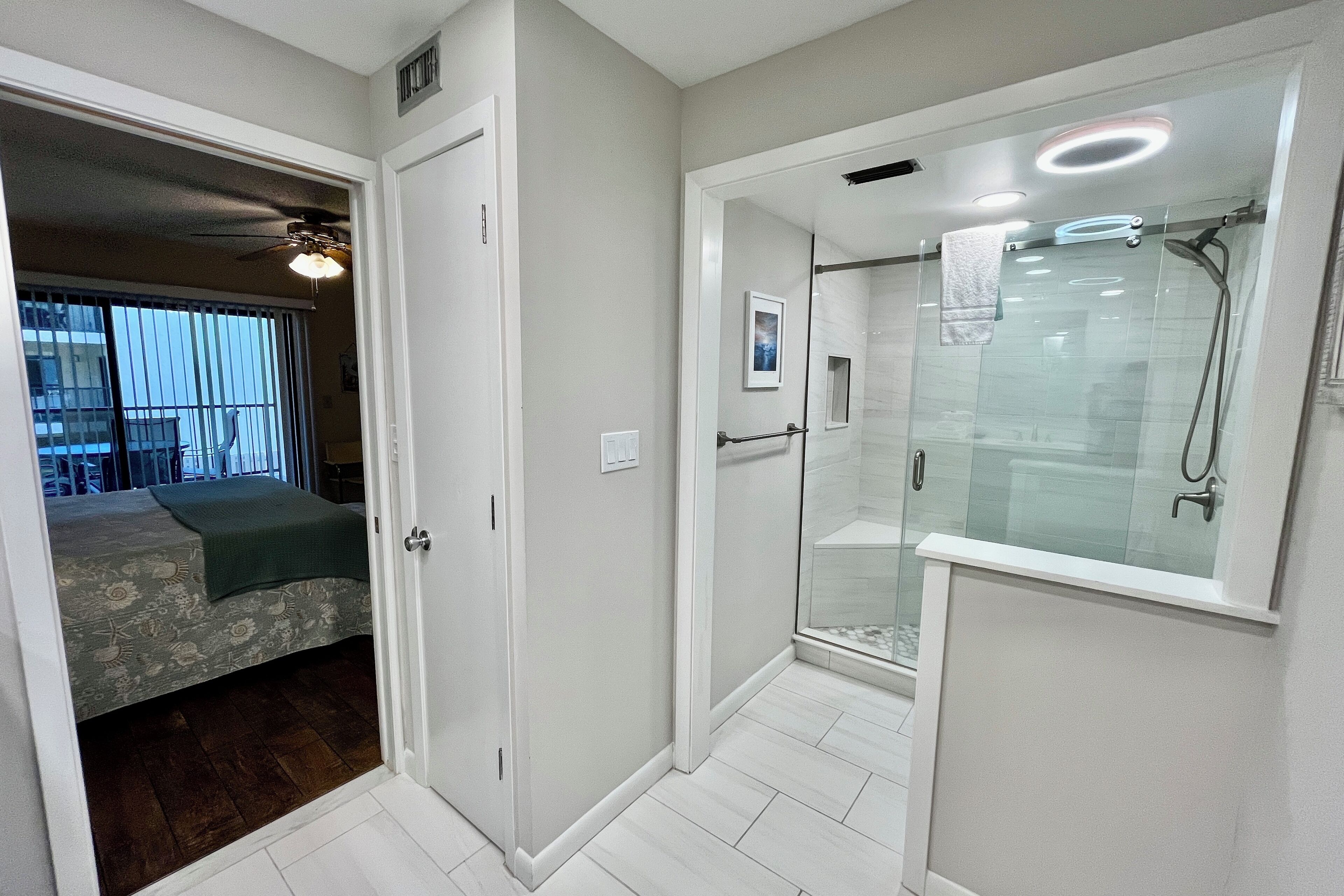 Un baño moderno con cabina de ducha de vidrio, un perchero visible y una puerta que da a un dormitorio con cama y ventana.