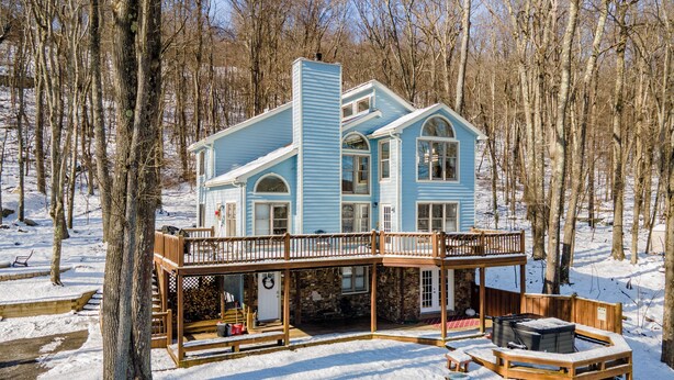 Casa de dos pisos con terraza de madera y cimientos de piedra, rodeada de un paisaje nevado y árboles.