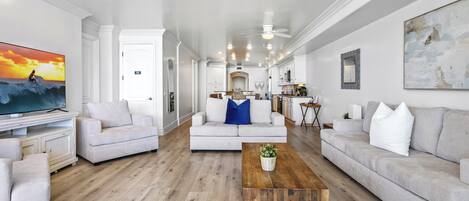 A spacious living room with a large flat-screen TV, two light-colored sofas, a wooden coffee table, and a kitchen area in the background.