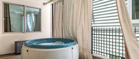 A hot tub in a room with a large window, a curtain, and a balcony with a railing.