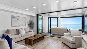 Ett rymligt vardagsrum med ett stort fönster som ger utsikt över havet, ett träbord och en beige soffa.