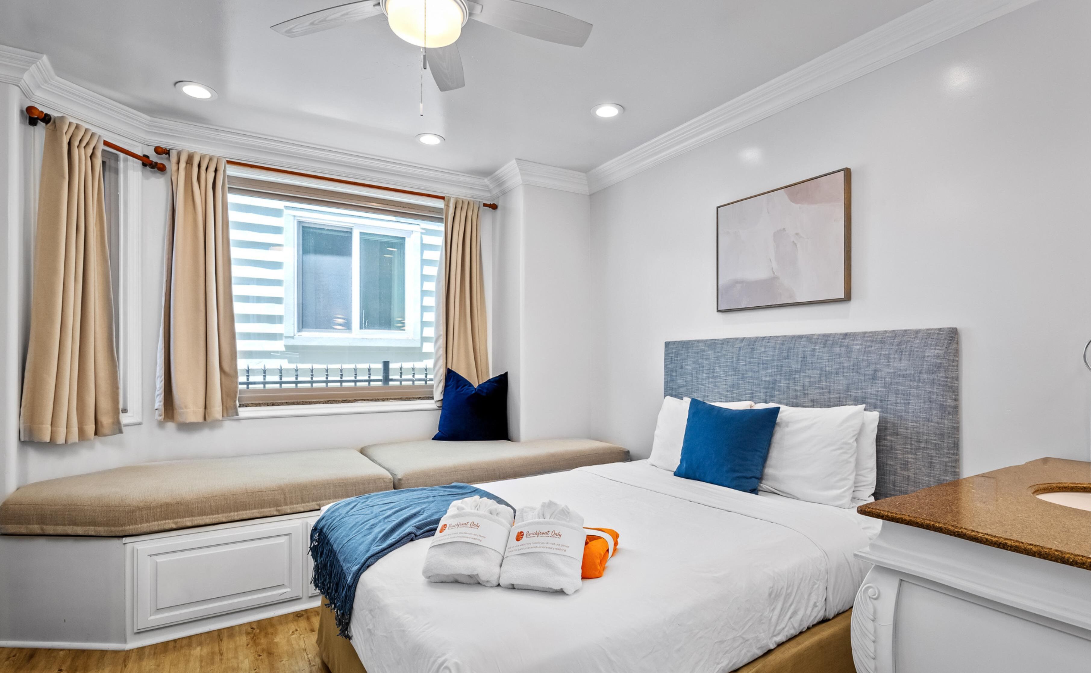 A hotel room with a bed, a bench, a window with curtains, a ceiling fan, a painting, and a bathroom counter.