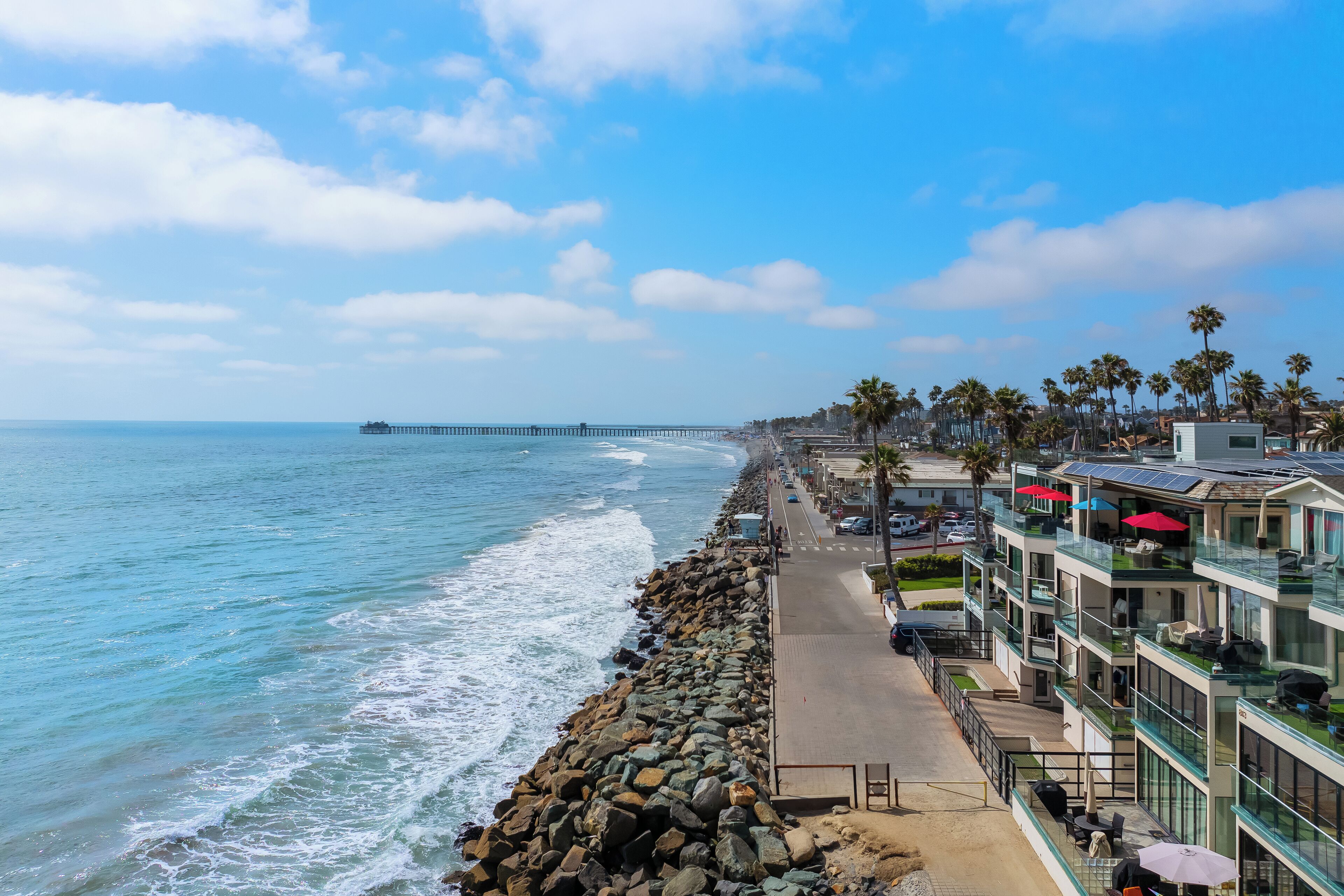 A coastal hotel with a rocky shoreline, a pier in the distance, and a row of palm trees.