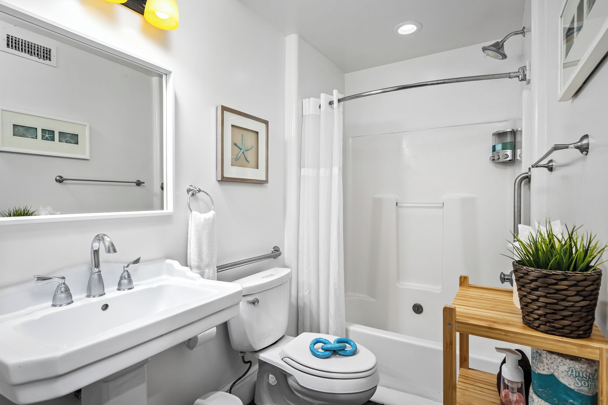 A modern bathroom with a white sink, toilet, and shower. There is a framed picture on the wall and a small plant on a wooden table.
