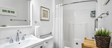 A modern bathroom with a white sink, toilet, and shower. There is a framed picture on the wall and a small plant on a wooden table.