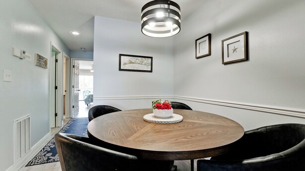 A dining area with a round wooden table, black chairs, and a white vase with red flowers. There are framed pictures on the wall and a ceiling light fixture.