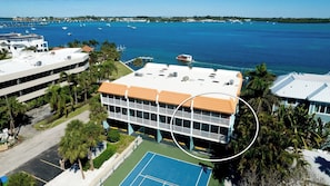 A multi-story building with a flat roof, surrounded by a tennis court and palm trees. - Pelican Cove 12 -  Anna Maria Island - Across the street from the beach- Condo  2 Bedroom /2 Bath bay view with a community pool,  maximum occupancy of 6 people. (Bradenton Beach)