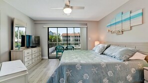 Een slaapkamer met een bed, een televisie, een dressoir, een balkon met stoelen en uitzicht op gebouwen.