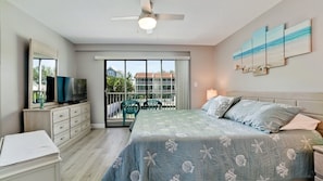 A bedroom with a bed, a TV, a dresser, a balcony with chairs, and a view of buildings. - Pelican Cove 12 -  Anna Maria Island - Across the street from the beach- Condo  2 Bedroom /2 Bath bay view with a community pool,  maximum occupancy of 6 people. (Bradenton Beach)