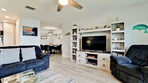 A modern living room with a dark blue sofa, a television on a wooden stand, and a dining area in the background.