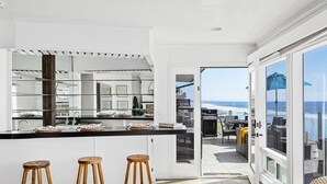 Eine moderne Küche mit einer großen Kochinsel, drei hölzernen Barhocker und Blick auf das Meer durch eine Glastür.
