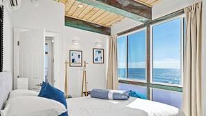 Ein Schlafzimmer mit großem Fenster und Meerblick, einem Bett mit weißer und blauer Bettwäsche sowie zwei gerahmten Kunstwerken an der Wand.