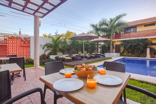Un área de comedor junto a la piscina con mesas, sillas y sombrillas.