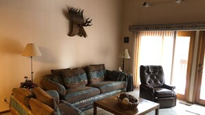 Sala de estar con sofá, sillón y mesa de centro. Se observa un ciervo montado en la pared como decoración y una ventana con cortinas.