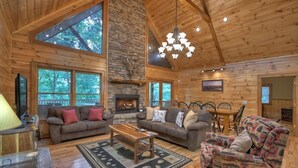 Un intérieur de cabane spacieux, doté d’une cheminée en pierre, de meubles en bois et de grandes fenêtres offrant une vue sur la forêt environnante.