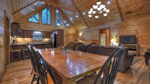 Une cabane en bois spacieuse, dotée d’une grande table à manger, de chaises, d’un canapé et d’un téléviseur.