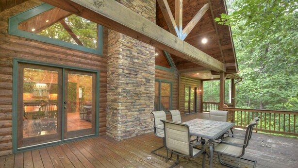 Une terrasse en bois avec un mur en pierre, des sièges d’extérieur et de grandes fenêtres donnant sur une forêt.