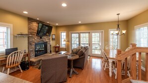 A spacious living room with a stone fireplace, a television mounted on the wall, and a dining area with wooden chairs and a table. - Swim, Ski & Scenic Lake Views by Deep Creek Lake (MCHENRY)