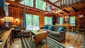 A spacious cabin with a large living area, wooden walls, and large windows offering a view of the forest. - Woodsy Dog Friendly Retreat w/ Lake Access (SWANTON)