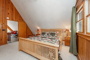 Een slaapkamer met een schuine balkenlaag, houten wanden, een bed met sprei, twee nachtkastjes met lampen en een raam met gordijnen.