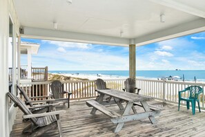Une terrasse en bois avec une table de pique-nique et des chaises, donnant sur une plage délimitée par une clôture et où se trouvent des baigneurs.