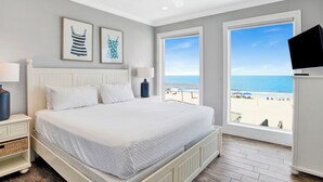 A bedroom with a bed, a ceiling fan, a TV, and a large window with a beach view.