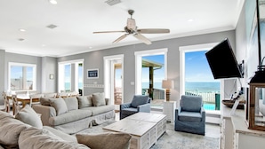 A spacious living room with a large sofa, two armchairs, a coffee table, and a ceiling fan. There is a TV mounted on the wall and a view of the ocean through large windows.