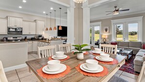 Una cocina amplia con armarios blancos, un comedor con mesa de madera preparada para cuatro personas, y una sala de estar con sofá y televisor.
