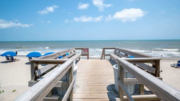 A wooden boardwalk leading to a beach with blue umbrellas and lounge chairs. - Ocean Dunes 2006 (Kure Beach)