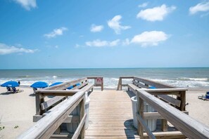 A wooden boardwalk leading to a beach with blue umbrellas and lounge chairs.