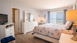 A bedroom with a bed, a TV, a dresser, a chair, and a window with curtains. - Ocean Dunes 2006 (Kure Beach)