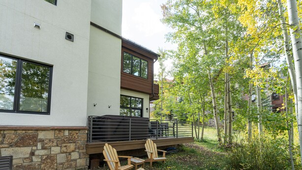 Une maison moderne avec des fondations en pierre, une terrasse en bois dotée de mobilier d’extérieur et une vue sur une zone boisée.