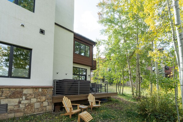 Ein modernes Haus mit Steinfundament, einer Holzterrasse mit Gartenmöbeln und Blick auf eine bewaldete Landschaft.