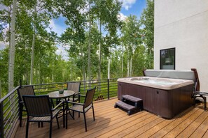Une terrasse en bois avec un jacuzzi, des chaises et une table, entourée d’arbres et d’un bâtiment.