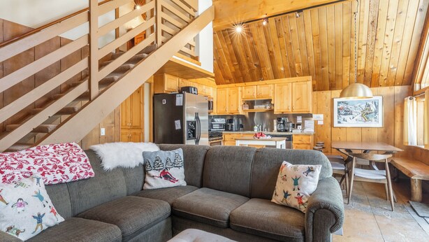 Een gezellig cabin interieur met een houten trap, een zithoek met een bank en kussens, een keuken met houten kastjes en een eettafel.
