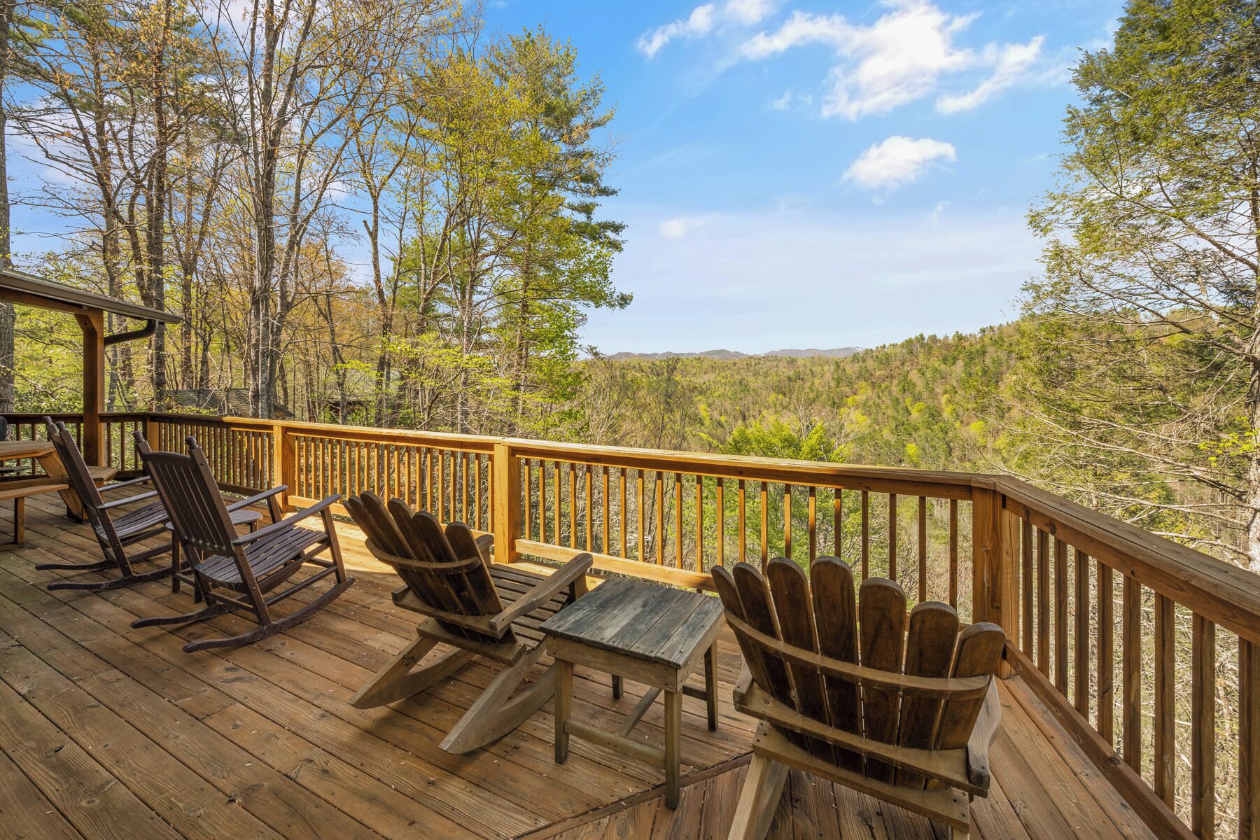 A wooden deck with Adirondack chairs and a table, overlooking a forested area.