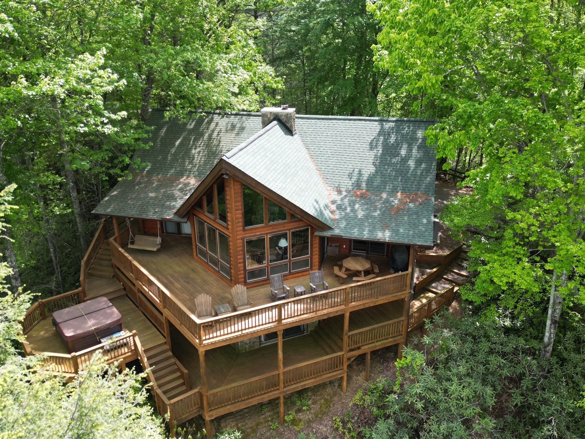 A cabin with a large deck, multiple windows, and a covered outdoor seating area surrounded by dense forest.