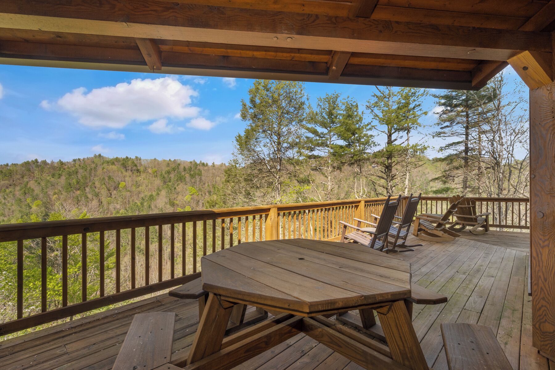 A wooden deck with a picnic table and Adirondack chairs, overlooking a forested hillside.