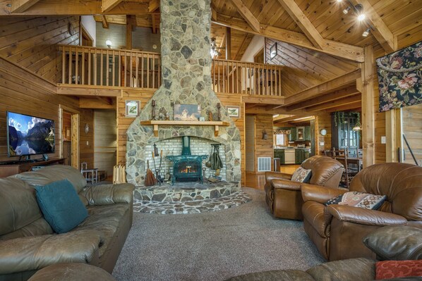 A cozy cabin interior with a stone fireplace, wooden beams, a television, leather sofas, and a kitchen in the background.