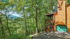 Una cabaña de madera con balcón, rodeada de un bosque denso y una montaña al fondo.