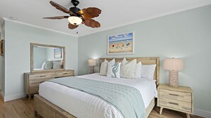 Een slaapkamer met een bed, nachtkastje, kapstok, plafondventilator en een schilderij aan de muur.
