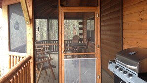 Une cabane en bois dotée d’un porche couvert, d’un barbecue et d’une porte moustiquaire donnant sur un espace extérieur aménagé pour s’asseoir.