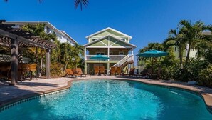 Ein Swimmingpool mit Liegestühlen und Sonnenschirmen, umgeben von Palmen und einem Gebäude mit Balkon.